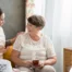 Smiling caregiver with her hand on an older woman’s shoulder as they share tea on the couch, showing home care vs assisted living