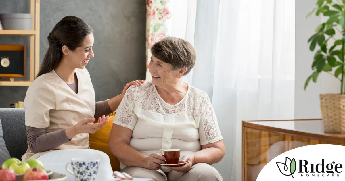 Smiling caregiver with her hand on an older woman’s shoulder as they share tea on the couch, showing home care vs assisted living